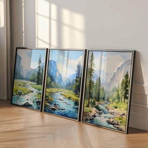 May include: Three framed landscape art prints featuring a river flowing through a valley with mountains and trees. The artwork has a watercolor style with blue, green, and brown tones. The frames are black with silver trim.