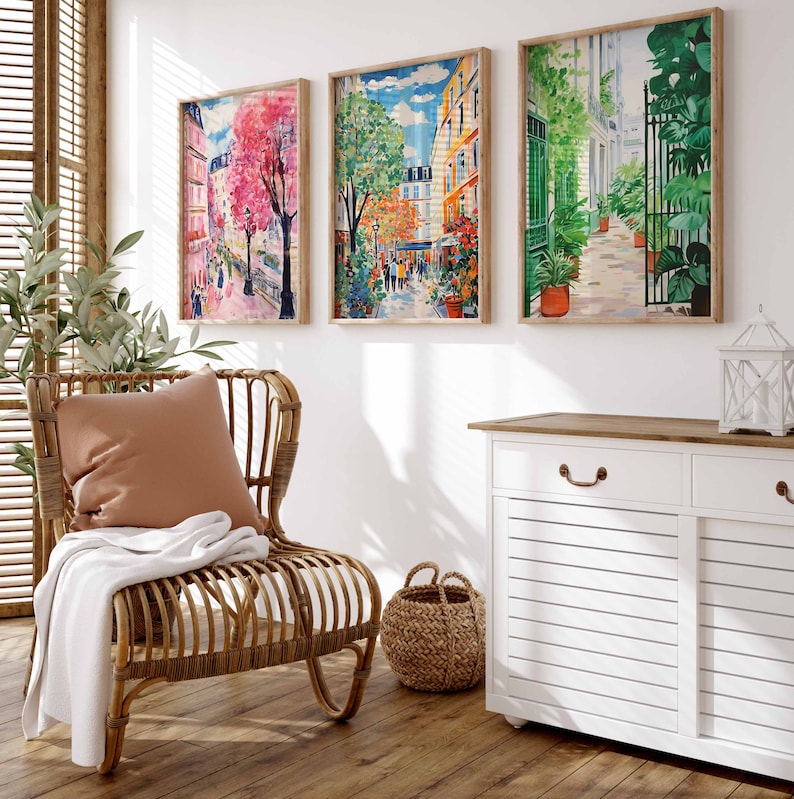 K&ouml;nnte beinhalten: Drei gerahmte Bilder mit bunten Stra&szlig;enszenen in Paris, Frankreich. Die Bilder h&auml;ngen an einer wei&szlig;en Wand &uuml;ber einem wei&szlig;en Schrank mit einer Holzplatte. Ein Korbstuhl mit einem braunen Kissen und einer wei&szlig;en Decke steht im Vordergrund.