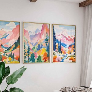 May include: Three framed landscape paintings with gold frames. The artwork features colorful mountain scenes with pink, blue, and green hues. The paintings are displayed on a white wall, with a plant in the foreground.
