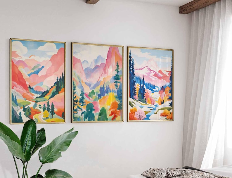 May include: Three framed landscape paintings with gold frames. The artwork features colorful mountain scenes with pink, blue, and green hues. The paintings are displayed on a white wall, with a plant in the foreground.