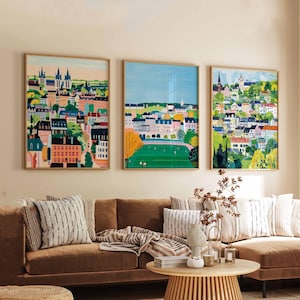 Könnte beinhalten: Drei gerahmte Illustrationen von bunten Stadtlandschaften mit Gebäuden, Bäumen und einem grünen Feld. Die Illustrationen hängen an einer Wand über einem braunen Sofa mit gestreiften Kissen und einer Wolldecke.