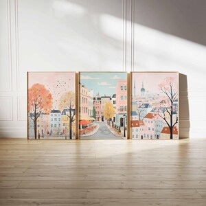 May include: A set of three framed prints depicting a city street scene. The first print shows a street with buildings on either side, a cafe with red umbrellas, and a tree with pink blossoms. The second print shows a street with buildings on either side, a cafe with red umbrellas, and a tree with pink blossoms. The third print shows a city skyline with a church steeple and a tree with pink blossoms.