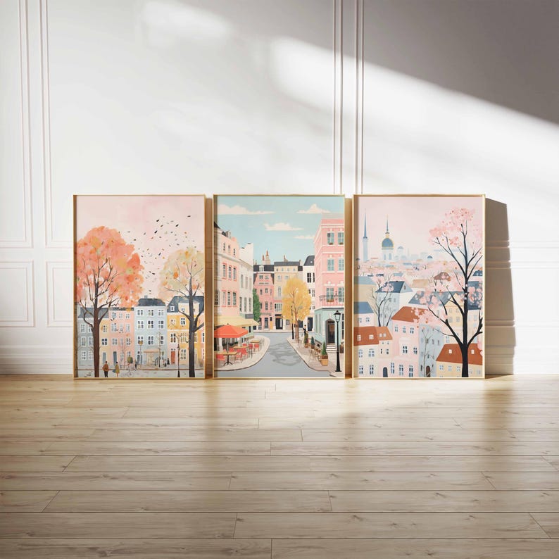 May include: A set of three framed prints depicting a city street scene. The first print shows a street with buildings on either side, a cafe with red umbrellas, and a tree with pink blossoms. The second print shows a street with buildings on either side, a cafe with red umbrellas, and a tree with pink blossoms. The third print shows a city skyline with a church steeple and a tree with pink blossoms.