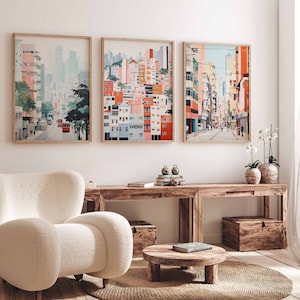 May include: Three framed watercolor paintings of cityscapes in pink, blue, green, and yellow hues. The paintings are hung on a white wall above a wooden table with a white armchair, a wooden coffee table, and two potted plants.