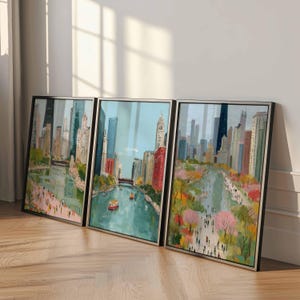 May include: Set of three framed cityscape art prints. The artwork depicts a river scene with buildings, boats, and people. The color palette includes blues, greens, pinks, and browns. The frames are black.