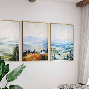May include: Three framed landscape art prints. Each print features a watercolor painting of a mountain scene with trees and a house. The frames are gold-colored, and the artwork is displayed on a white wall.