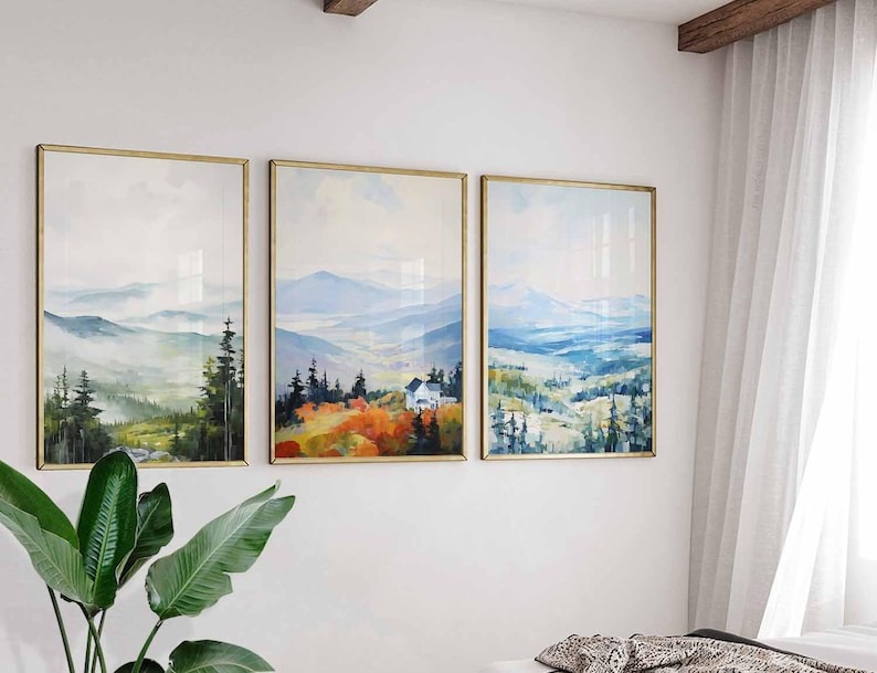 May include: Three framed landscape art prints. Each print features a watercolor painting of a mountain scene with trees and a house. The frames are gold-colored, and the artwork is displayed on a white wall.