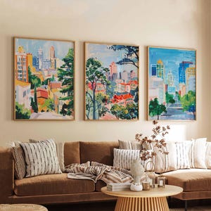 May include: Three framed paintings of cityscapes with colorful buildings and trees. The paintings are hung on a light beige wall above a brown couch with throw pillows and a blanket.