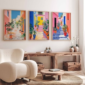 May include: Three colorful framed prints of a street scene with potted plants and buildings. The prints are hung on a white wall above a wooden table and a white armchair.