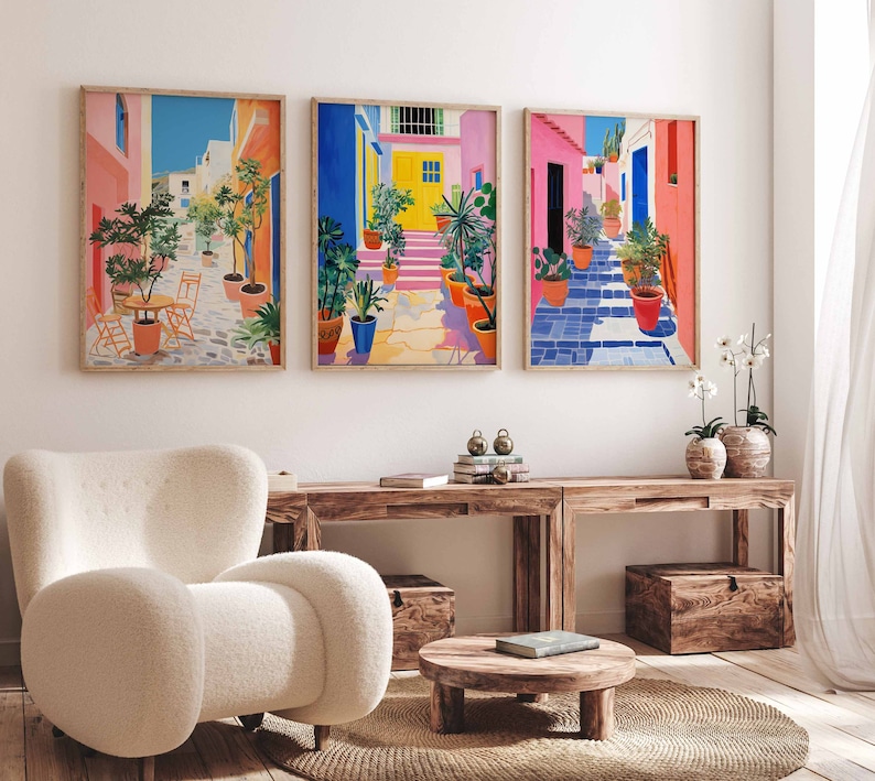 May include: Three colorful framed prints of a street scene with potted plants and buildings. The prints are hung on a white wall above a wooden table and a white armchair.