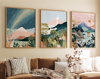 May include: Three framed landscape art prints. The artwork features mountain scenes with pink, green, and blue hues. The frames are a light wood color. The art is displayed above a brown sofa with striped pillows.