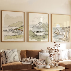 May include: Three framed landscape art prints. Each print features a pen and ink style drawing of a coastal scene with mountains, water, and buildings. The frames are a light wood color, and the art has a neutral color palette.