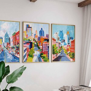 May include: Three framed cityscape paintings with gold frames hang on a white wall. The artwork depicts colorful city streets with buildings and blue skies. The paintings are displayed above a bed and next to a large green plant.