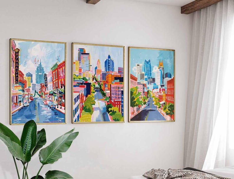 May include: Three framed cityscape paintings with gold frames hang on a white wall. The artwork depicts colorful city streets with buildings and blue skies. The paintings are displayed above a bed and next to a large green plant.