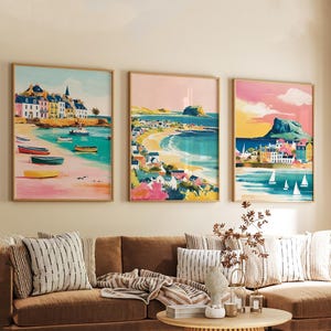 以下が含まれることがあります： 海岸の風景を描いた3つの額入りアートプリント。各プリントには、ビーチ、建物、ボート、カラフルな空が描かれています。アートワークは水彩画風で、ピンク、青、黄色などの暖色系の色使いです。プリントは茶色のソファの上に飾られています。