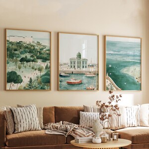 May include: Three framed watercolor prints depicting coastal scenes. The artwork features a park with trees, a harbor with boats, and a beach with ocean views. The frames are a light brown color, and the overall theme is a coastal landscape.