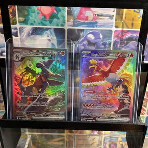 May include: Two Pokemon trading cards in protective sleeves. One card features Cynthia's Garchomp EX with the text "Corkscrew Dive" and "Draconic Buster". The other card shows Ethan's Ho-Oh EX with "Golden Flame" and "Shining Feathers". Both cards are displayed in a black card holder.