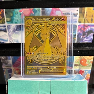May include: A gold Mega Charizard Y EX Pokémon card in a protective sleeve. The card features a detailed illustration of Charizard and the text "Explosion Y". The card is displayed on a light blue box.