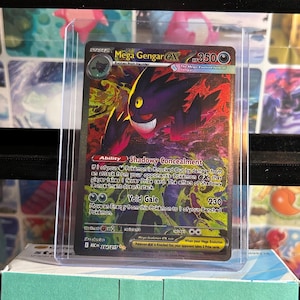 May include: A Mega Gengar EX Pokémon card in a protective sleeve. The card features a purple and black Gengar with a menacing grin, set against a fiery background. The card has the text "Mega Gengar EX" and "Void Gale".