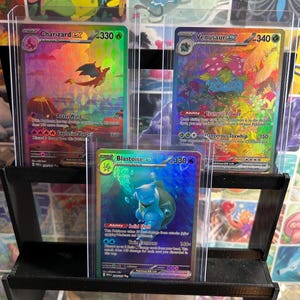 May include: Three holographic Pokémon trading cards displayed in protective cases. The cards feature Charizard, Venusaur, and Blastoise, each with unique artwork and game information. The cards are on a black tiered display.