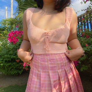 May include: A pink crocheted crop top with a lace-up detail at the front, worn with a pink and white plaid skirt.