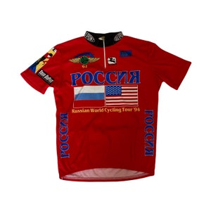 Peut inclure: Maillot de cyclisme rouge avec "РОССИЯ" en lettres bleues, avec les drapeaux russe et américain. Le maillot comprend le texte "Russian World Cycling Tour '94" et divers logos de sponsors. Col noir et fermeture éclair partielle.
