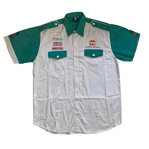 May include: A white short-sleeved button-down shirt with green sleeves and a red bull logo on the front. The shirt has several patches, including "Petronas Malaysia", "Credit Suisse", and "Petronas".
