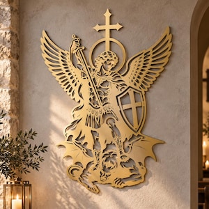 May include: A gold-coloured metal wall art piece depicting Saint Michael the Archangel with wings, a sword, and a shield, standing over a defeated figure. The intricate cutout design features a cross and halo above the central figure's head.
