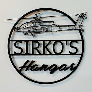 May include: Black metal wall art featuring a helicopter design. The helicopter is positioned above a circular frame with the text "SIRKO'S Hangar" cut out. The artwork is mounted on a white wall.
