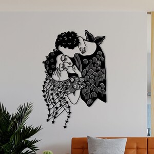 Op de afbeelding: Zwart metalen wandkunst die twee figuren in een omhelzing afbeeldt. Het kunstwerk heeft ingewikkelde uitgesneden details, waaronder vloeiend haar en kleding met patronen. De figuren staan tegen een witte muur in een woonkamer.