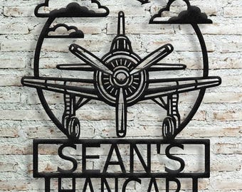 May include: Black metal airplane wall art with the text "SEAN'S HANGAR". The airplane is centered within a circular frame, with clouds and a bird silhouette above. The sign is mounted on a brick wall.