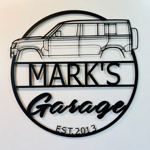 Puede incluir: Un letrero de metal negro con la silueta de un Land Rover Defender, el texto "MARK'S Garage" y el texto "EST. 2013".