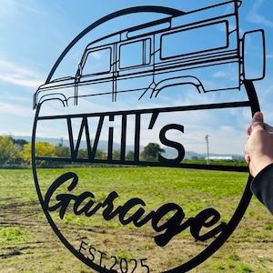 May include: Black metal sign featuring a vehicle outline within a circular frame. The sign reads "Will's Garage" with "EST. 2025" below. The background shows a green field and a blue sky.