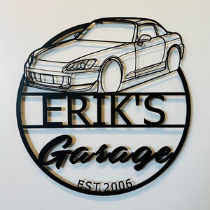 May include: Black metal sign with a silhouette of a Honda S2000 sports car, the text "Erik's Garage" and the date "EST. 2006".
