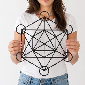 Op de afbeelding: Een zwartmetalen Metatrons Cube wandkunstwerk. Het geometrische ontwerp heeft onderling verbonden cirkels en lijnen, waardoor een symmetrisch patroon ontstaat. Het kunstwerk wordt vastgehouden door een persoon die een wit T-shirt en een blauwe spijkerbroek draagt.