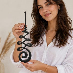 May include: Black decorative wall art with a spiral and swirl design. The art piece has a vertical orientation and is held in front of a neutral background. The design is made of a solid black material.