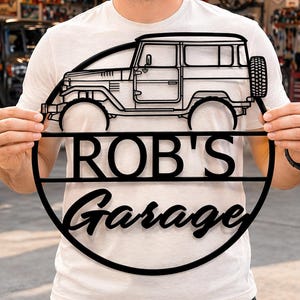 Op de afbeelding: Zwart metalen bord met een gedetailleerde omtrek van een vintage SUV, met de tekst "ROB'S Garage" in een cirkelvormig ontwerp. Het bord wordt voor een garage gehouden, wat wijst op een thema van autodecoratie.