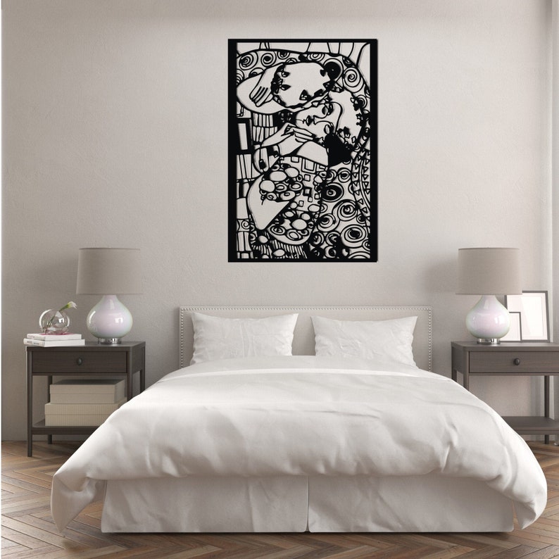 Op de afbeelding: Zwart-witte wandkunst met een gestileerd paar dat elkaar omarmt. Het kunstwerk is ingelijst in een zwart frame en hangt aan een witte muur boven een wit bed met witte beddengoed.