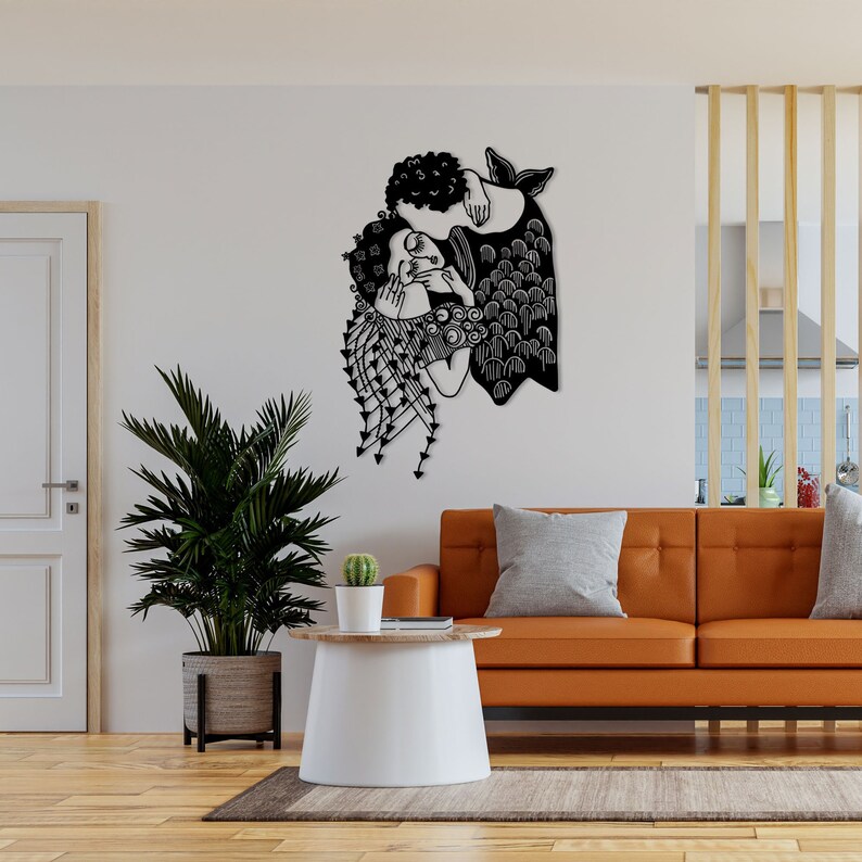 Op de afbeelding: Zwart metalen wandkunst die twee figuren in een omhelzing afbeeldt. Het kunstwerk heeft ingewikkelde uitgesneden details, waaronder vloeiend haar en kleding met patronen. De figuren staan tegen een witte muur in een woonkamer.
