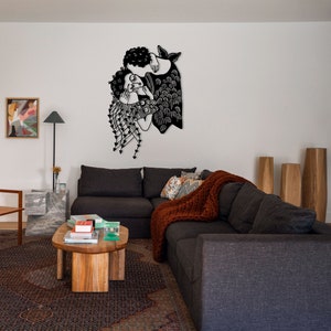 Op de afbeelding: Zwart metalen wandkunst die twee figuren in een omhelzing afbeeldt. Het kunstwerk heeft gedetailleerde kenmerken, waaronder krullend haar en vloeiende kleding. Het is gemonteerd op een witte muur boven een donkergrijze hoekbank.