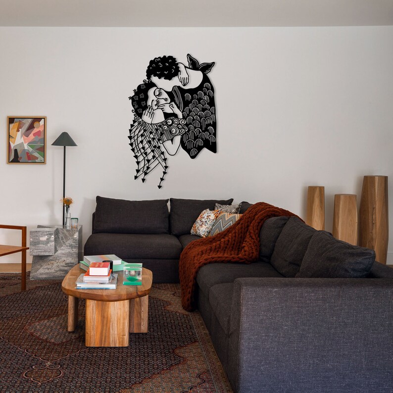 Op de afbeelding: Zwart metalen wandkunst die twee figuren in een omhelzing afbeeldt. Het kunstwerk heeft gedetailleerde kenmerken, waaronder krullend haar en vloeiende kleding. Het is gemonteerd op een witte muur boven een donkergrijze hoekbank.