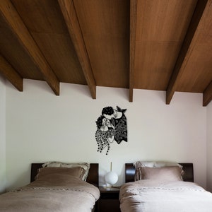 Op de afbeelding: Zwart-witte wandkunst met twee figuren die elkaar omarmen. Het kunstwerk heeft ingewikkelde patronen en details, waarbij een figuur op een zeemeermin lijkt. De kunst is gemonteerd op een witte muur boven twee bedden met bruin beddengoed.