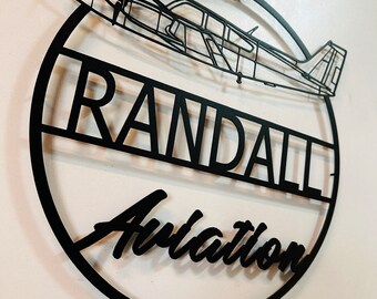 May include: Black metal wall art featuring an airplane silhouette above the words "RANDALL Aviation" within a circular frame. The design is cut from metal and mounted on a white wall.