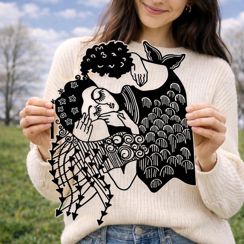 Op de afbeelding: Een zwart-wit zeemeerminkunstwerk wordt door een persoon vastgehouden. Het kunstwerk toont twee zeemeerminnen die elkaar omarmen, met ingewikkelde details in hun haar, staarten en kleding. Het stuk is uit &eacute;&eacute;n vel gesneden, waardoor een uniek silhouet ontstaat.