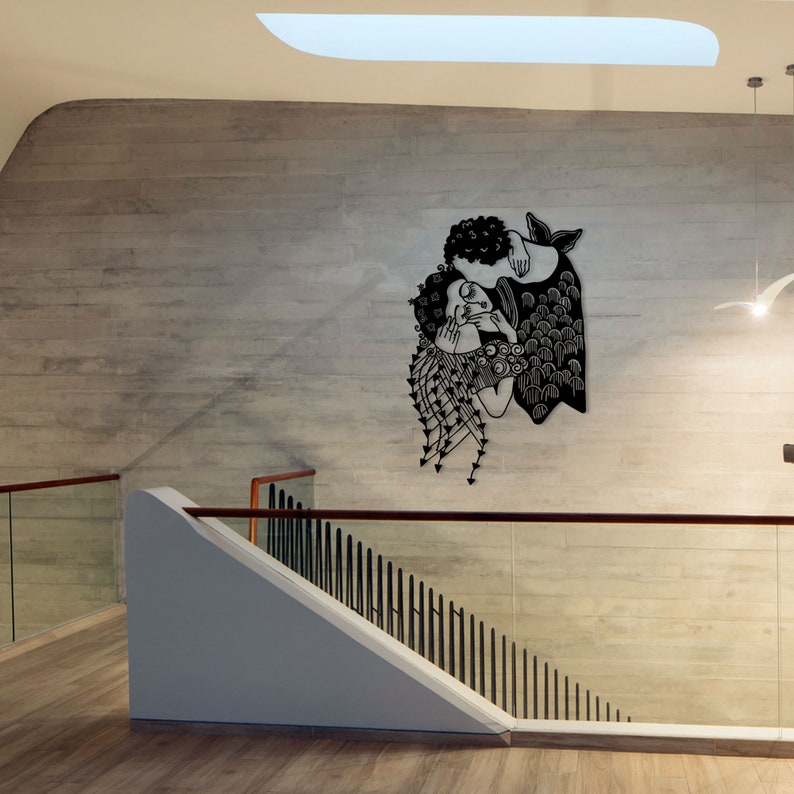 Op de afbeelding: Een zwart metalen wandkunstwerk dat twee figuren omarmend afbeeldt. Het kunstwerk is bevestigd aan een lichtgrijze betonnen muur, met een trap en een leuning op de voorgrond. Het stuk heeft ingewikkelde details en een moderne esthetiek.