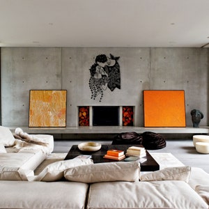 Op de afbeelding: Een moderne woonkamer met een betonnen muur. Een zwart metalen wandkunstwerk met twee figuren die elkaar omarmen, is aan de muur bevestigd. De kamer heeft een beige bank, een donkere houten salontafel en abstracte schilderijen in oranje en geel.
