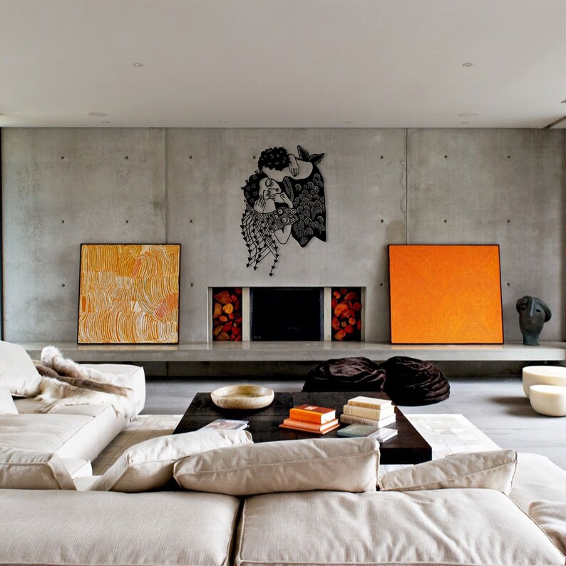 Op de afbeelding: Een moderne woonkamer met een betonnen muur. Een zwart metalen wandkunstwerk met twee figuren die elkaar omarmen, is aan de muur bevestigd. De kamer heeft een beige bank, een donkere houten salontafel en abstracte schilderijen in oranje en geel.