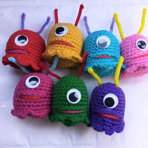 Könnte beinhalten: Sieben gehäkelte Plüschtiere in Form von Geistern in verschiedenen Farben, darunter Rot, Gelb, Blau, Rosa, Lila, Grün und Pink. Jedes Spielzeug hat ein großes Auge und einen kleinen roten Mund.