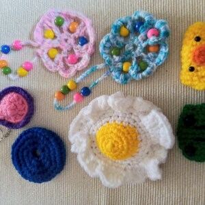 May include: A collection of crocheted items, including a pink and white flower with colourful beads, a blue and white flower with colourful beads, a pink and purple heart, a blue doughnut, a white and yellow egg, a yellow chick, and a green frog.