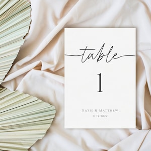 May include: White table number card with black calligraphy that says "table 1" and the names Katie & Matthew and the date 17.12.2022.